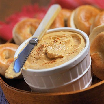 Some pumpkin spread in a white container wth a knife and some bagels.