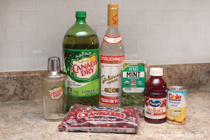 Bottle of vodka, Canada Dry, cranberry juice, cranberries. pineapples and a shaker on the counter.