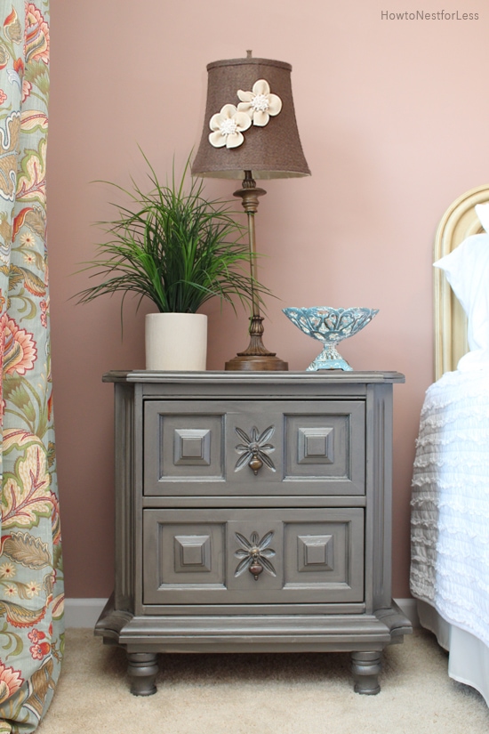 A gray nightstand beside the bed with a lamp, bowl and potted plant on it.