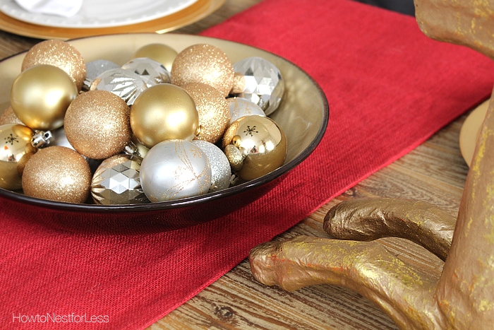 A bowl filled with gold and silver sparkly ornaments in the middle of the table on a red table runner.