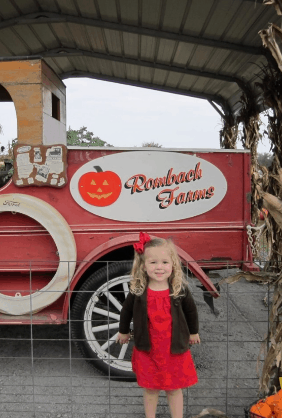 Blonde girl with a red bow in front of a red truck at the pumpkin patch.