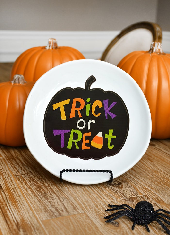 White plate with trick or treat in a black pumpkin on it, on a plate stand in front of pumpkins.