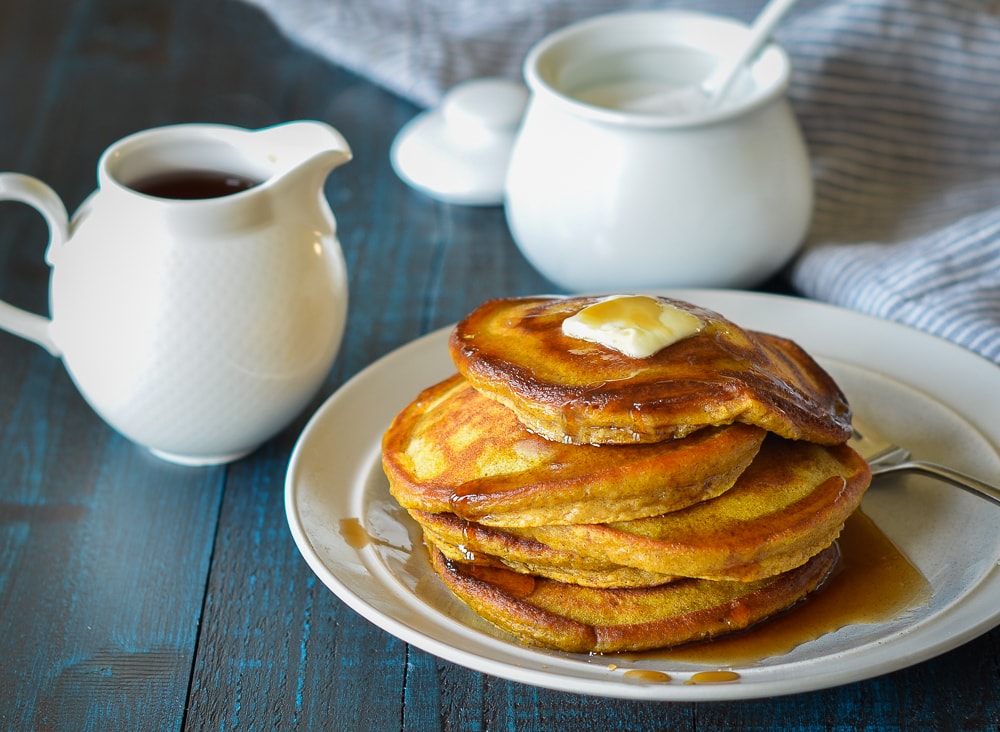 Pumpkin pancakes on a white plate with syrup on top on a blue table.