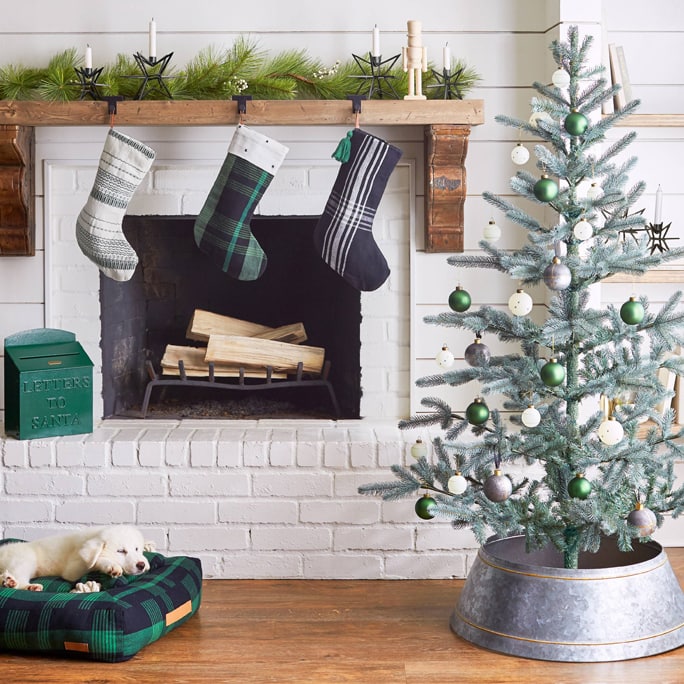 An aluminum Christmas tree collar around the tree, and a white washed fireplace with stockings in hung up in front of it.