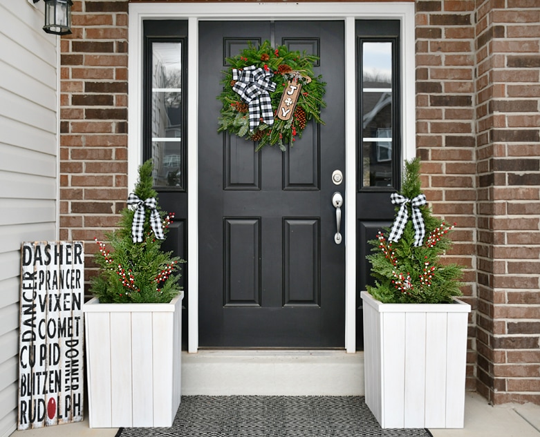A decorated front porch with a wreath on the door and two mini trees decorated beside the door.