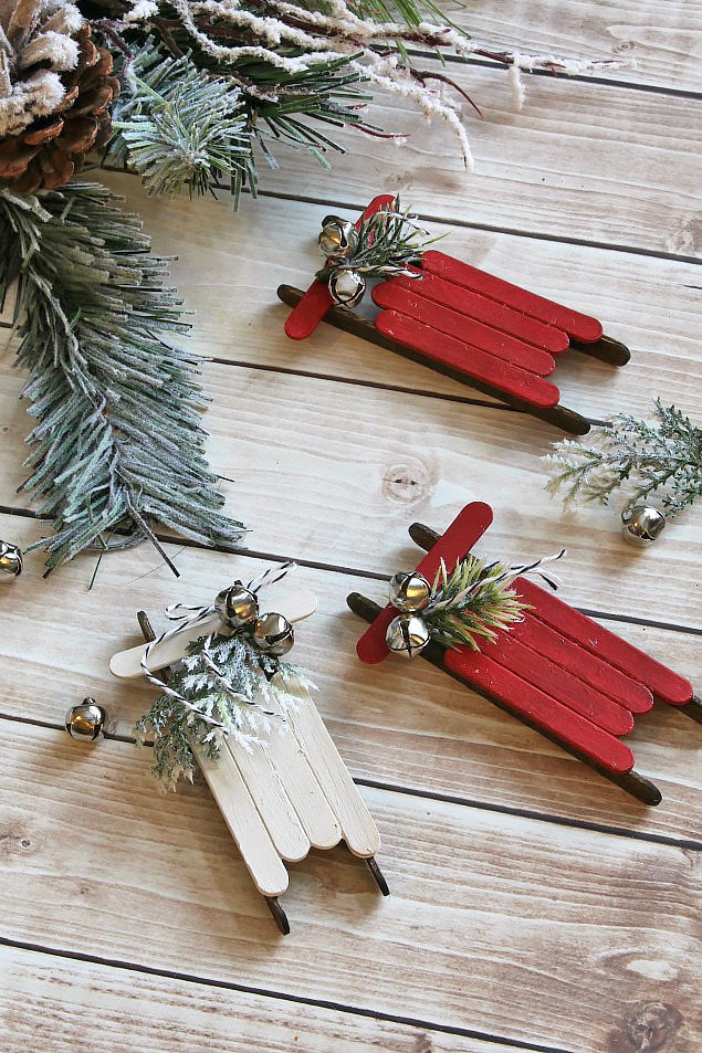 Popsicle stick sleighs in red and white with bells lying on the ground under the tree.