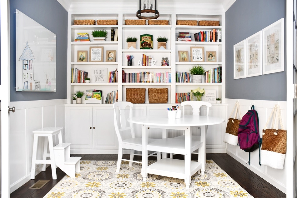 Room with blue walls, white board and batten on the lower part of the walls and white built-in bookshelves and cabinets on the far wall. A whiteboard is hung on the left wall and art prints are hung on the right wall. Backpacks and bags hang on hooks on the board and batten. A white table with white chairs in the middle of the room on a beige rug.