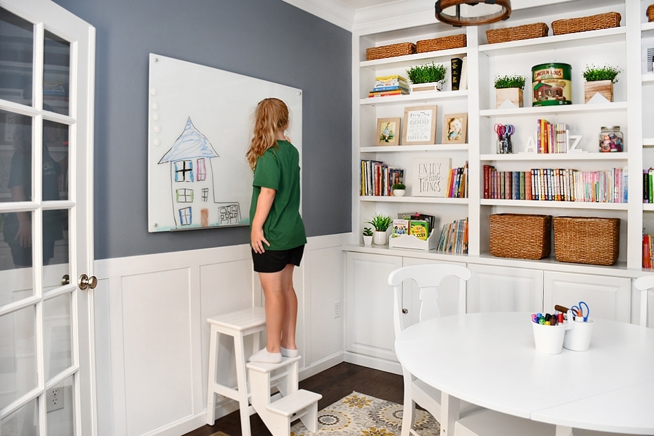 A young girl wearing a green shirt and dark shorts stands on a white step stool. She's writing on a whiteboard that's mounted on the wall.