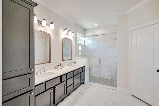 Bathroom painted Eider White. A large walk-in shower with large marble tiles is at the far end of the bathroom. A double vanity with cabinetry lines a wall.