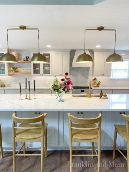 Large kitchen with large kitchen island with white marble countertops. Kitchen cabinets and kitchen island are painted Eider White by Sherwin Williams. Brass pulls are on the cabinets. Two brass lighting fixtures hang above the kitchen island.
